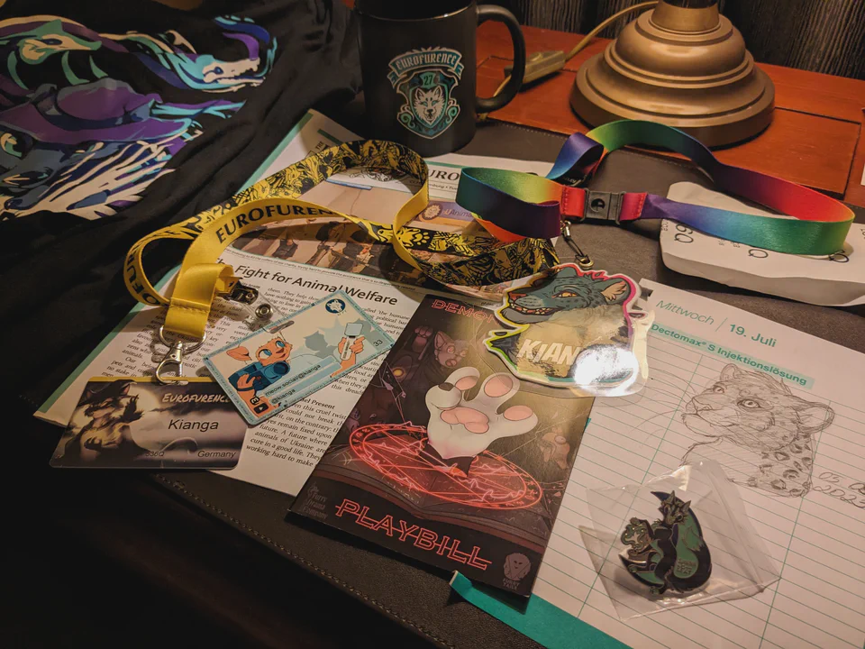 Lots of stuff lying on my hotel desk again: the official EF27 con shirt, an EF27 mug, my con, video creator, and fursona badges, the Daily Eurofurence newspaper, the playbill for the Demonormicon by Furry Tails Theater, a paper with a bust sketch of my jaguar fursona made by my friend MadKakerlaken, and an EF27 sponsor pin.