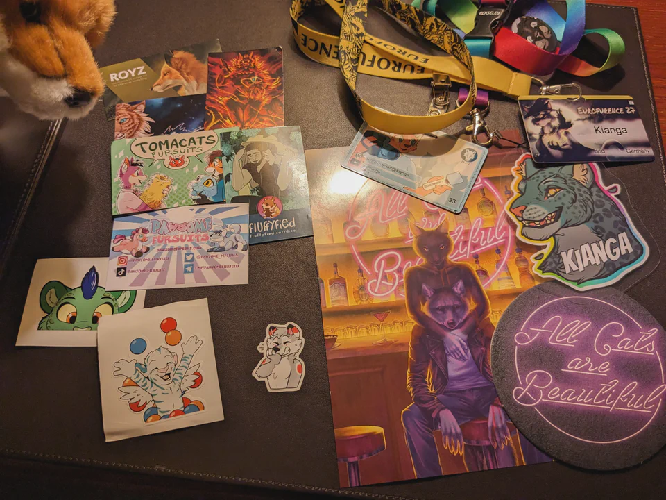 Photo of various things on my hotel desk: my con badge, fursona badge, media badge, colorful business cards from various artists, a couple of stickers, a playbill and coaster from the pawpet show “All Cats Are Beautiful”, and the snout of a red fox plush peeking into the picture from the top left.
