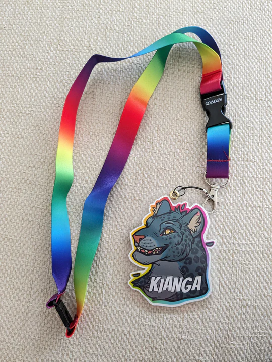 Photo of my profile icon as a laminated convention badge. The icon is a bust of a smiling anthropomorphic jaguar with dark gray fur and golden eyes, with a bright rainbow-colored outline, and the badge is cut like the shape of the icon, with the name KIANGA in big white letters on the bottom part.  It's attached to a lanyard made of the same bright rainbow colors as the outline of the icon.
