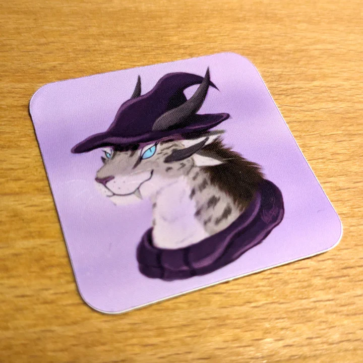 Close-up photo of a printed vinyl sticker lying on a wooden surface. The sticker is a bust of a female charr mesmer with beige fur, a dark brown mane, and light blue eyes, against a soft light purple background. She has a confident smile and is wearing a dark purple wizard hat, with her two horns poking through it.