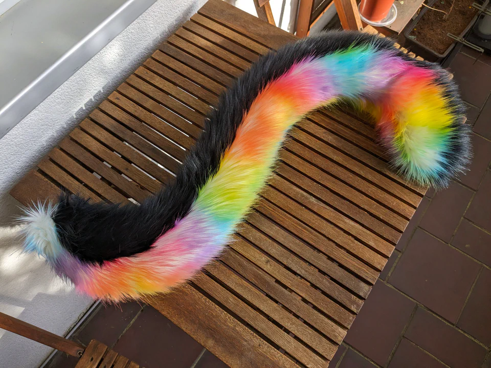 Photo of a fursuit tail on our balcony table. It has rainbow-colored fur on the top side and black fur on the bottom.
