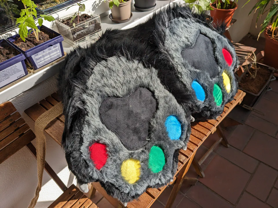 Photo of two large digitigrade fursuit foot paws lying on a wooden table, bottoms up to show the gray soles and colorful paw pads (red, yellow, green, blue, and black).
