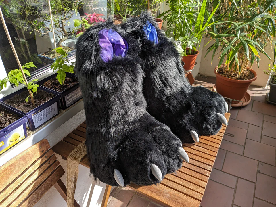 Photo of two very large digitigrade fursuit paws standing on a wooden table. They're made of long black fur, and have gray claws and lining with a purple galaxy pattern.
