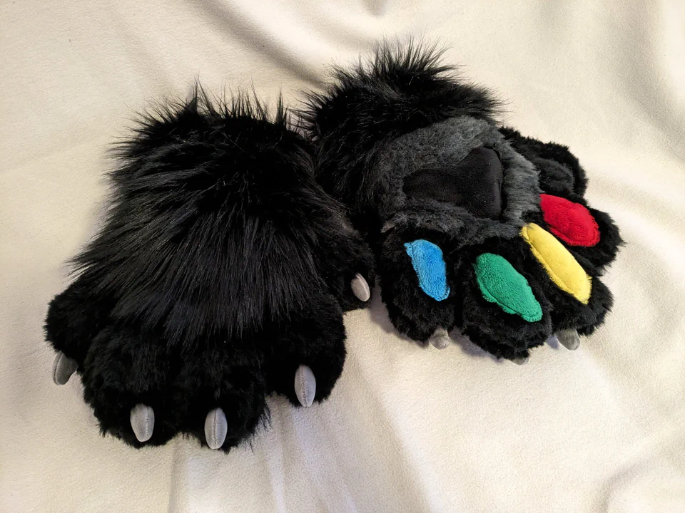 Photo of two fursuit hand paws lying on soft white fabric: they have long black fur on the top, shorter charcoal fur on the palm, and short black fur and gray claws on the fingers. The palm and thumb paw pads are black, the other finger paw pads are red, yellow, green, and blue.
