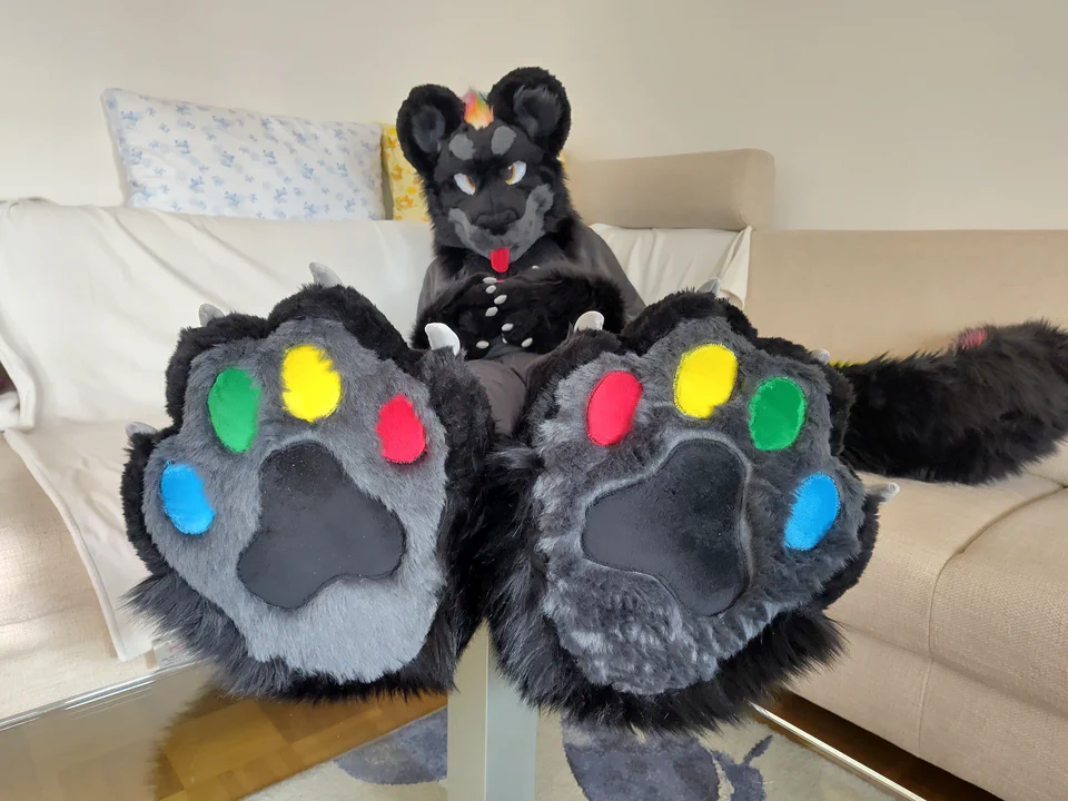 Photo of me wearing my fursuit, sitting on a white couch, legs up showing the colorful paw pads.
