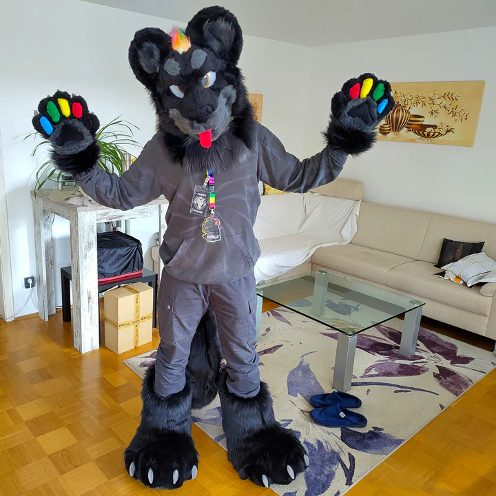 Photo of me wearing my black jaguar partial fursuit: It has black fur, amber eyes, a grey muzzle with soft teeth and a long red tongue, and a rainbow-colored mane that flows between the ears down the back, and big fluffy digitigrade foot paws.
